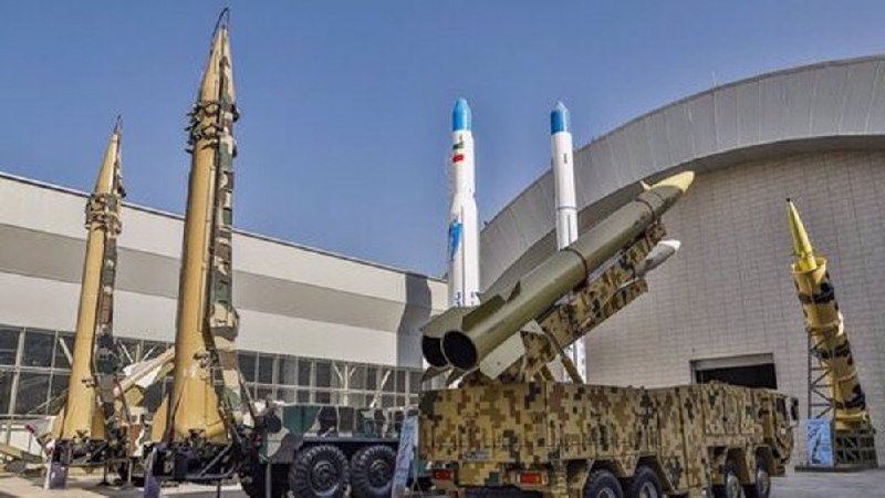 Several large missiles and rockets are displayed on mobile launchers outside a modern building under a clear blue sky.