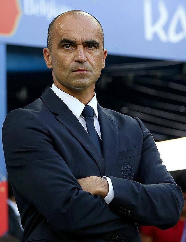 A bald man in a dark suit and navy tie stands with crossed arms, with a crest visible on his jacket pocket.