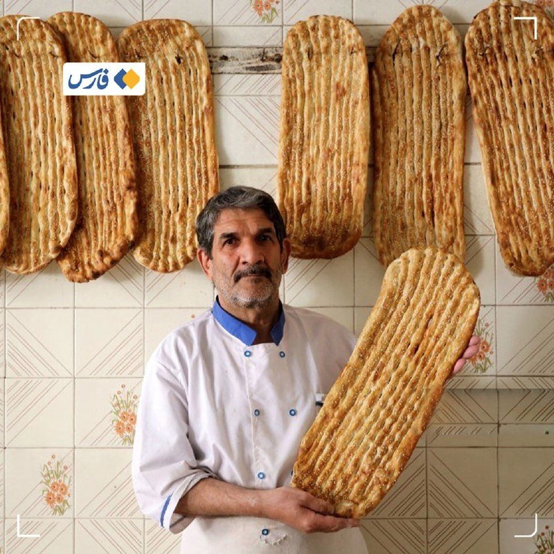 A man with a mustache, wearing a white shirt, holds a large oval flatbread against a tiled wall displaying several similar flatbreads.