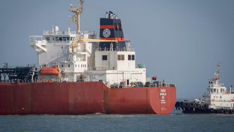 A large red-hulled cargo ship with a white superstructure is accompanied by a small tugboat in calm blue waters under a clear sky.