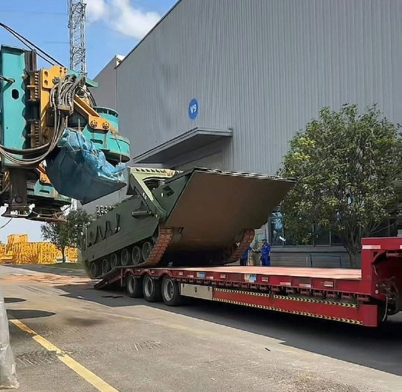 A crane lowers an olive-green military bridge-laying vehicle onto a red flatbed trailer outside a large industrial facility.