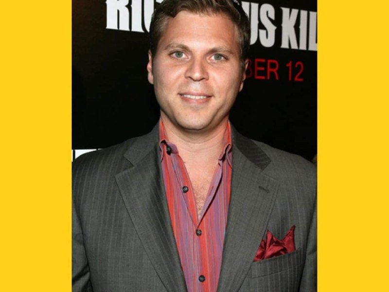 A smiling man in a gray pinstripe suit and striped shirt poses in front of a promotional backdrop displaying partial text including "KIL" and "BER 12."