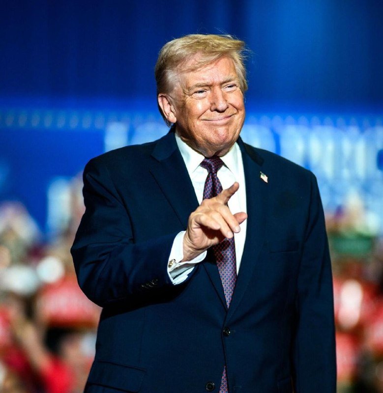 A blonde-haired man in a dark suit and purple tie points forward while smiling on a stage with a blue-lit background and an audience visible below.