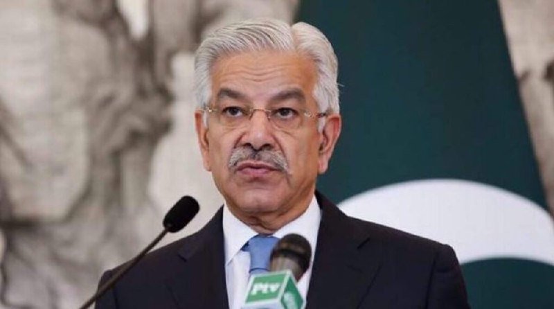 An older man with gray hair, a mustache, and glasses speaks at a podium with a PTV microphone, standing before a green and white flag backdrop.