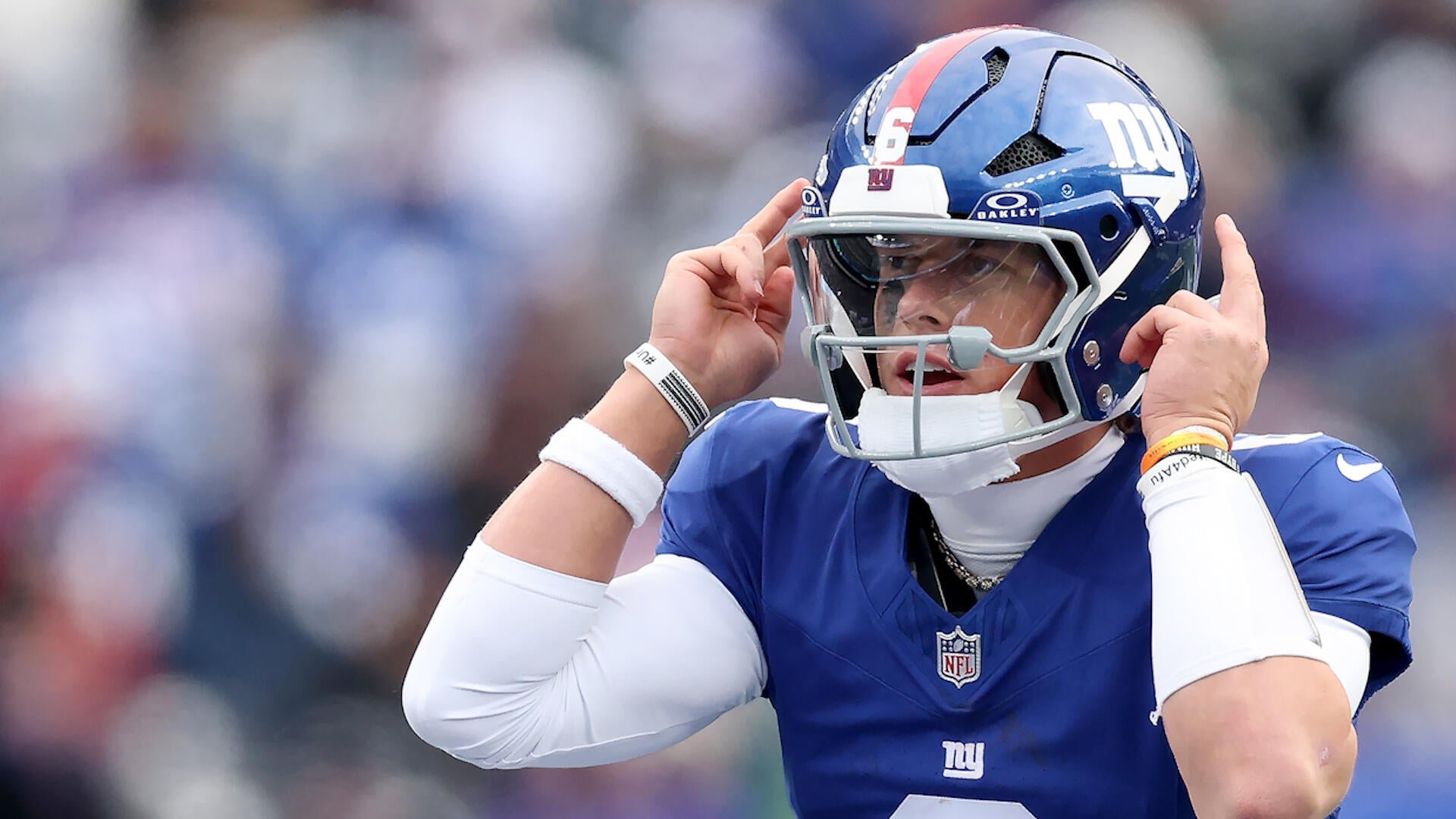 A football player in a blue New York Giants jersey and helmet adjusts his facemask with both hands on the field.
