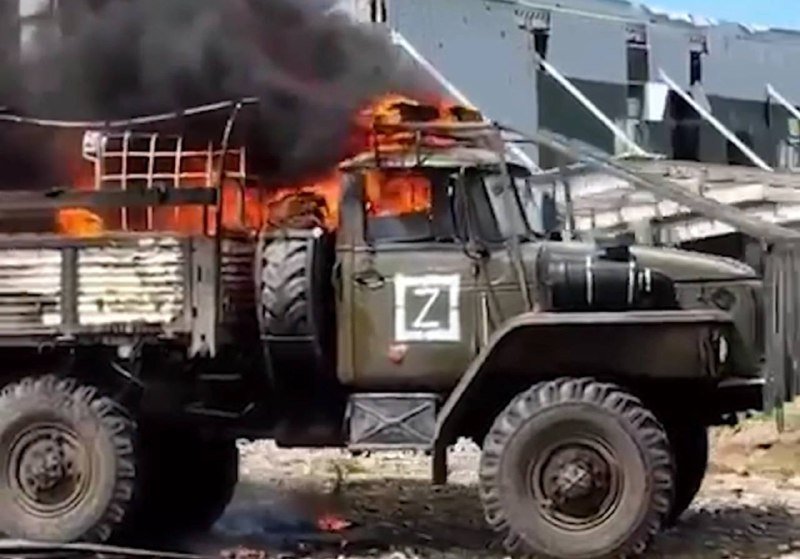 A military truck marked with a white "Z" symbol is engulfed in flames and emitting thick black smoke in front of a building.