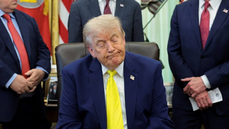 A man in a navy suit and yellow tie sits with pursed lips at a desk, flanked by three men in suits standing behind him near American flags.