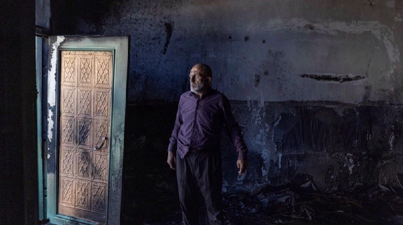 A man stands inside a burned, smoke-damaged room beside a charred wooden door, surrounded by debris and blackened walls.