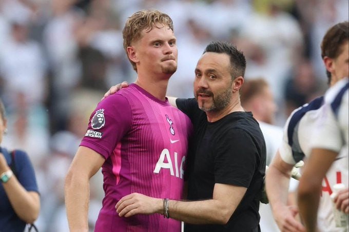 A Tottenham player in a pink away kit stands beside a man in a black shirt with an arm around the player's shoulder on a football pitch.