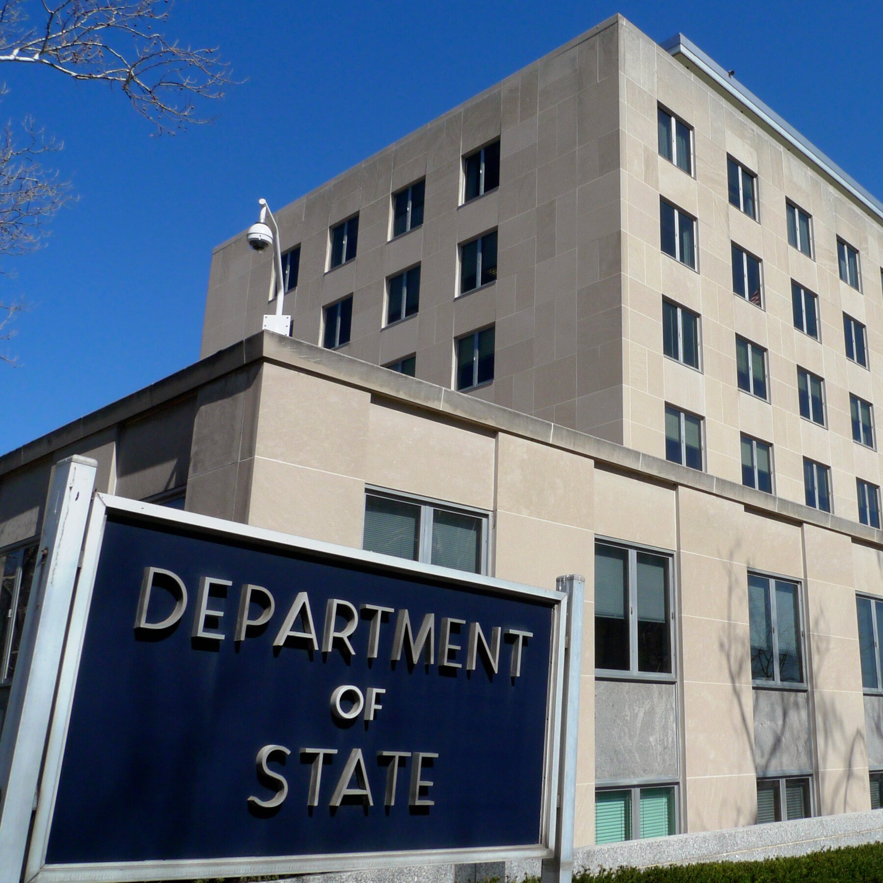 A multi-story stone government building stands under a clear blue sky, with a prominent blue sign reading "DEPARTMENT OF STATE" displayed in the foreground.