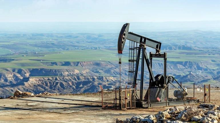 An oil pumpjack operates on a hilltop overlooking a vast expanse of green fields and rugged terrain under a clear sky.