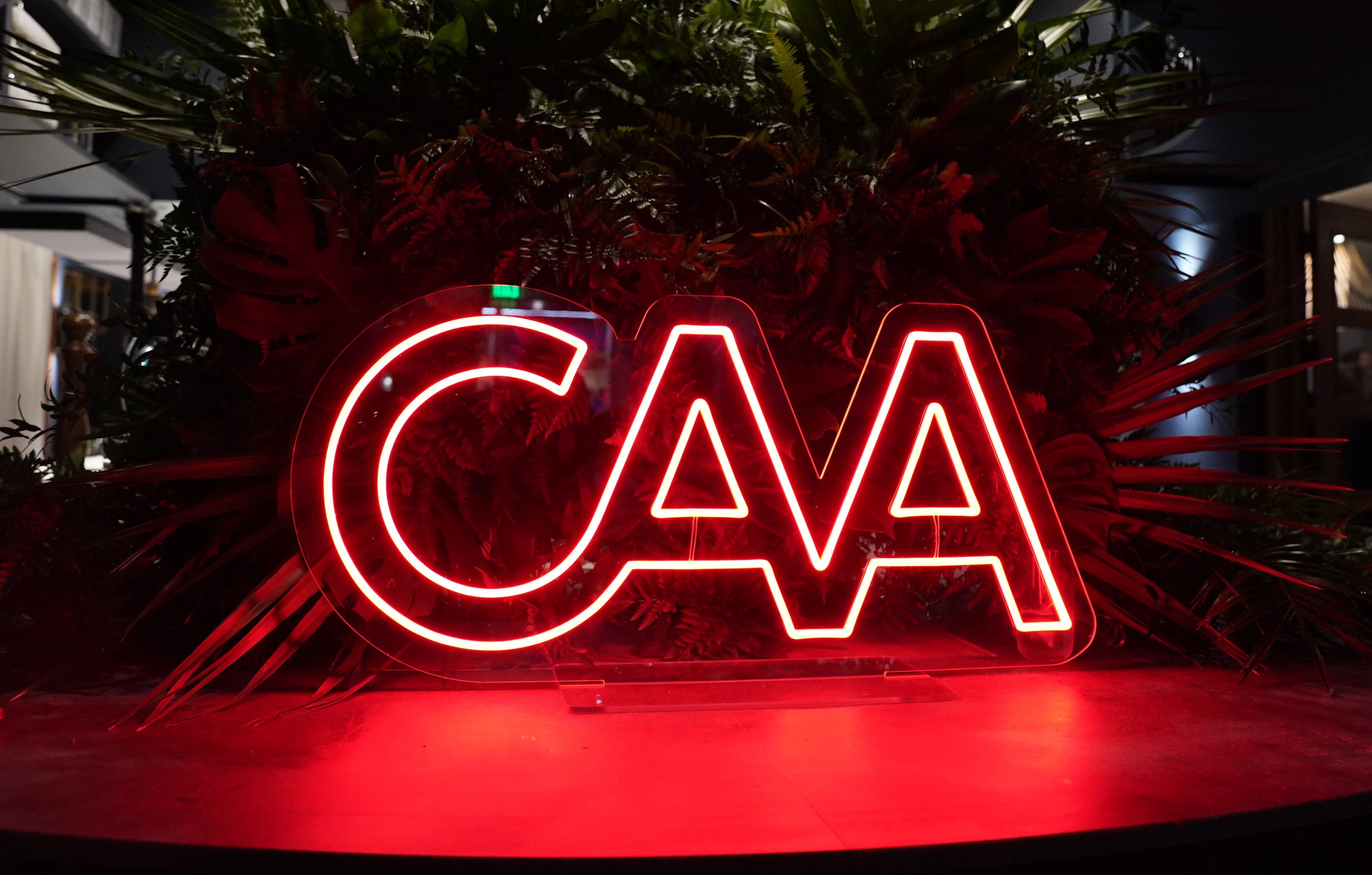A red neon-lit "CMA" sign displayed on an acrylic base sits in front of a dense arrangement of tropical plants.