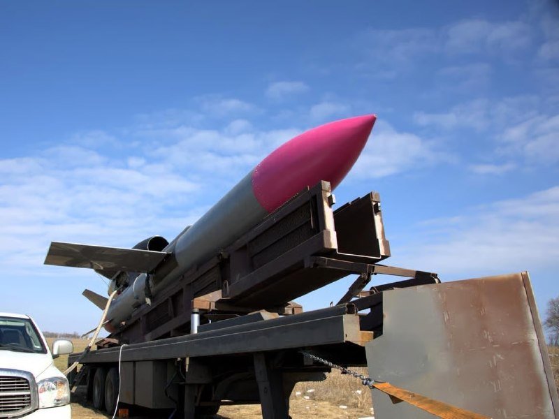 A large rocket with a bright pink nose cone sits mounted on a metal trailer attached to a white pickup truck in an open field under a blue sky.