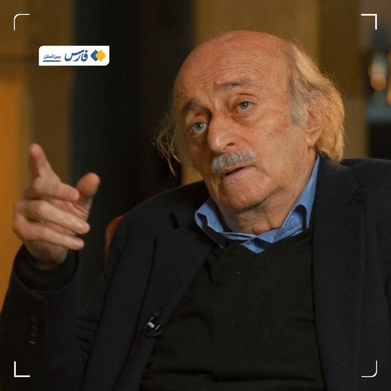 An elderly man with a mustache, wearing a dark jacket over a blue shirt, gestures with his right hand during what appears to be an interview, with a Persian-language logo in the corner.