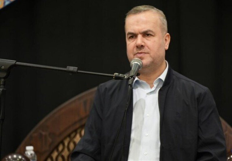 A man in a dark jacket and white shirt speaks into a microphone against a dark background.