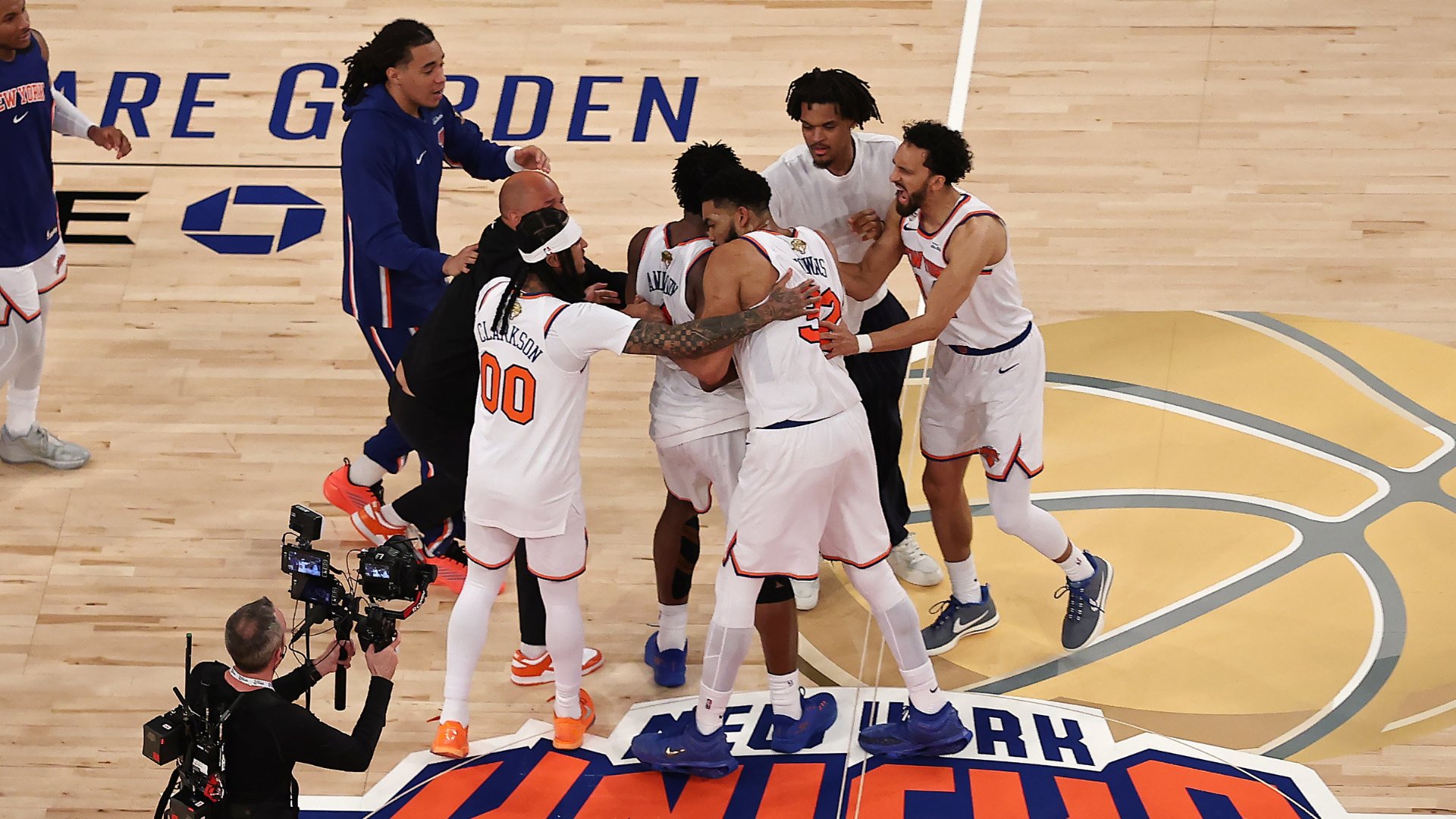 New York Knicks players in white uniforms embrace and celebrate on a basketball court while a cameraman films them from the sideline.