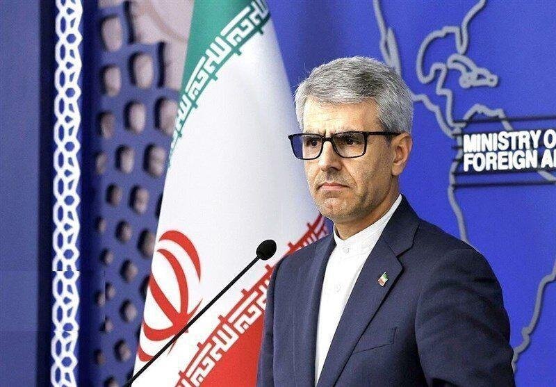 A gray-haired man in glasses and a dark suit stands at a microphone beside an Iranian flag, with a "Ministry of Foreign Affairs" backdrop behind him.