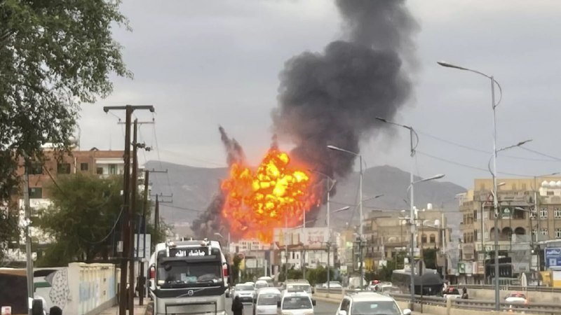 A large explosion with bright orange flames and thick black smoke rises above a city street, with vehicles and a truck visible below.