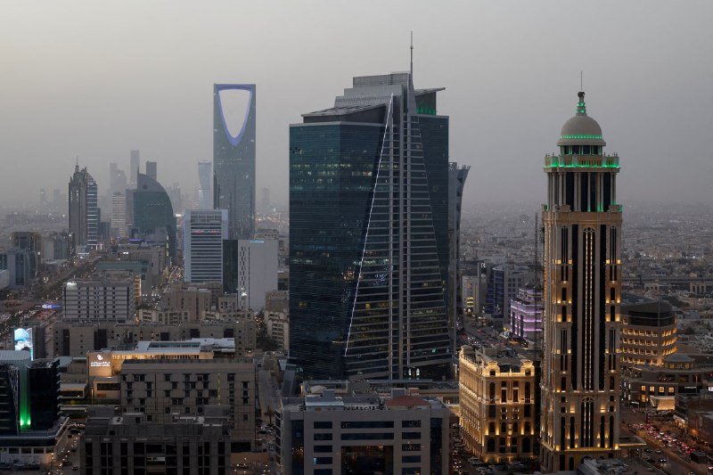 A hazy cityscape features a cluster of illuminated skyscrapers, including a tall building with a distinctive rectangular opening near its top and a domed tower glowing with green lights.