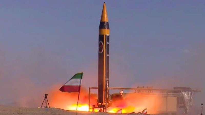 A large ballistic missile launches vertically from a desert site, with flames and smoke billowing at its base beside an Iranian flag.