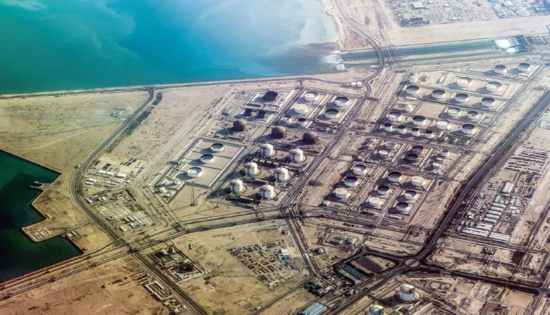 Aerial view of a large coastal industrial facility featuring numerous circular storage tanks and spherical containers along a turquoise shoreline.
