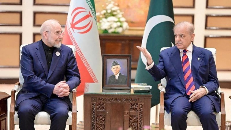 Two men in suits sit facing each other in a formal meeting, with Iranian and Pakistani flags behind them and a framed portrait displayed between them.