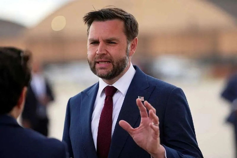 A bearded man in a dark suit and red tie speaks while gesturing with his hand, set against a blurred outdoor background.