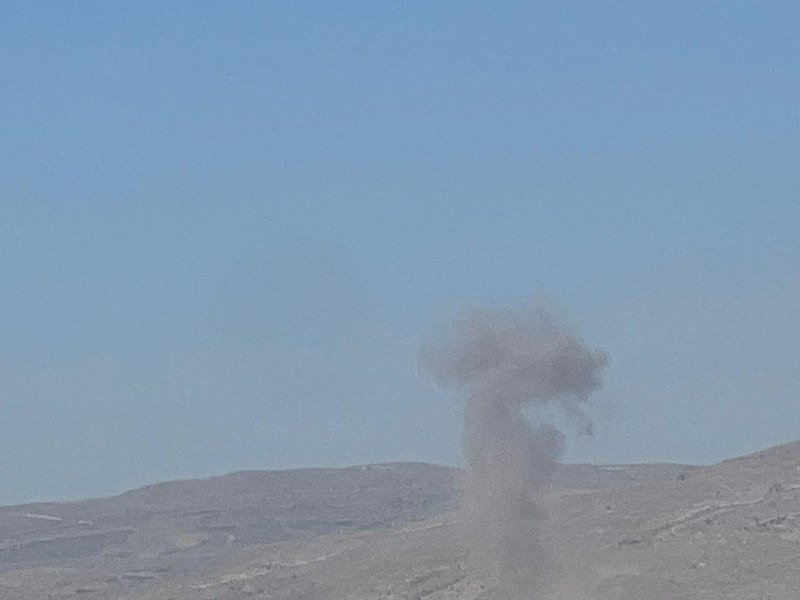 A plume of dark smoke rises above a barren, rocky hillside beneath a clear blue sky.