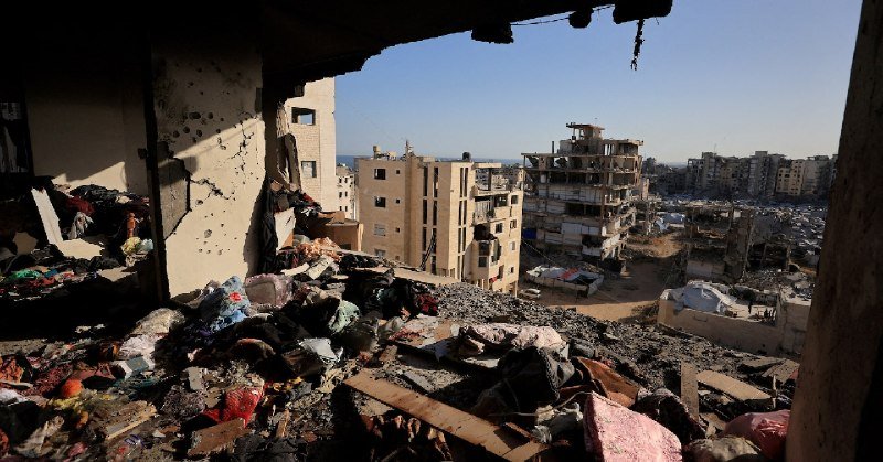 Rubble, debris, and scattered belongings fill a damaged building interior, overlooking several multi-story structures with visible destruction under a clear sky.