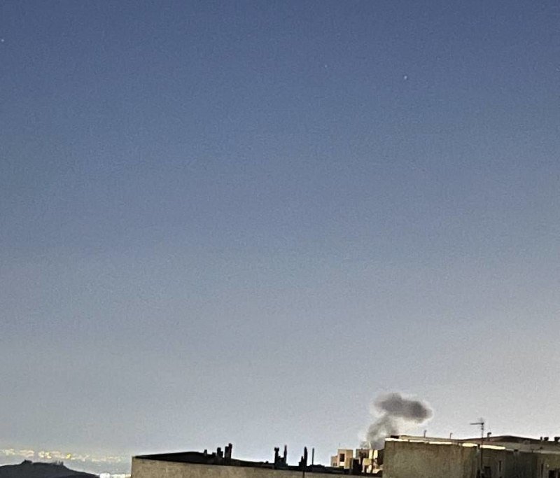 A plume of gray smoke rises above flat-roofed buildings against a pale blue sky with snow-capped mountains visible in the distance.