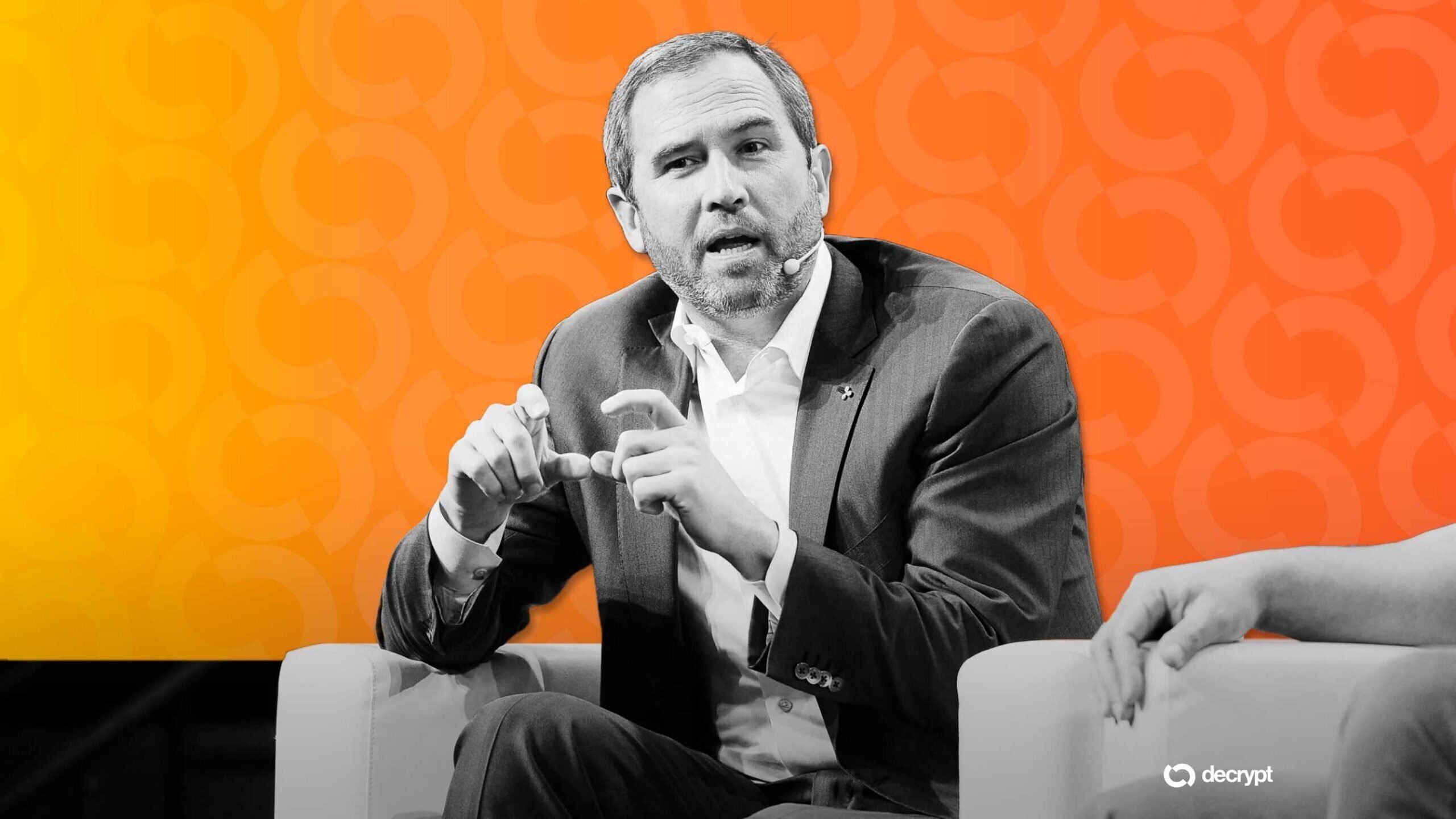 A gray-haired man in a suit gestures while speaking on stage with an orange backdrop, with the "decrypt" logo visible in the corner.