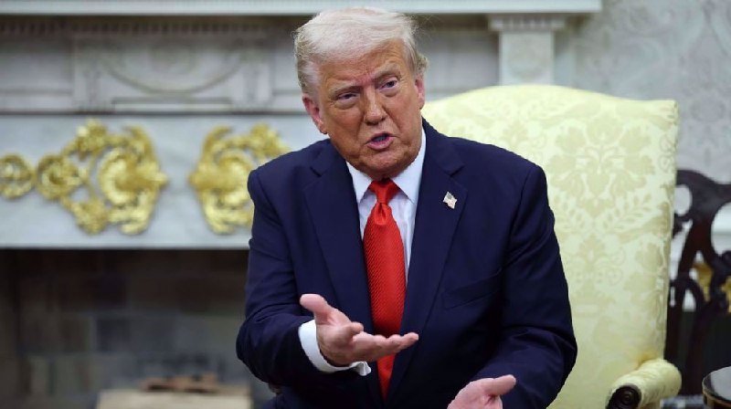 A man with blonde hair wearing a navy suit and red tie sits in a yellow-patterned chair, gesturing with his hands in front of an ornate marble fireplace.