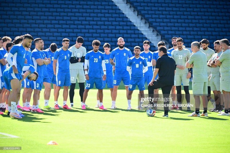 Soccer players in blue and light green uniforms gather on a grass field, listening to a coach in black pointing toward a ball at his feet.