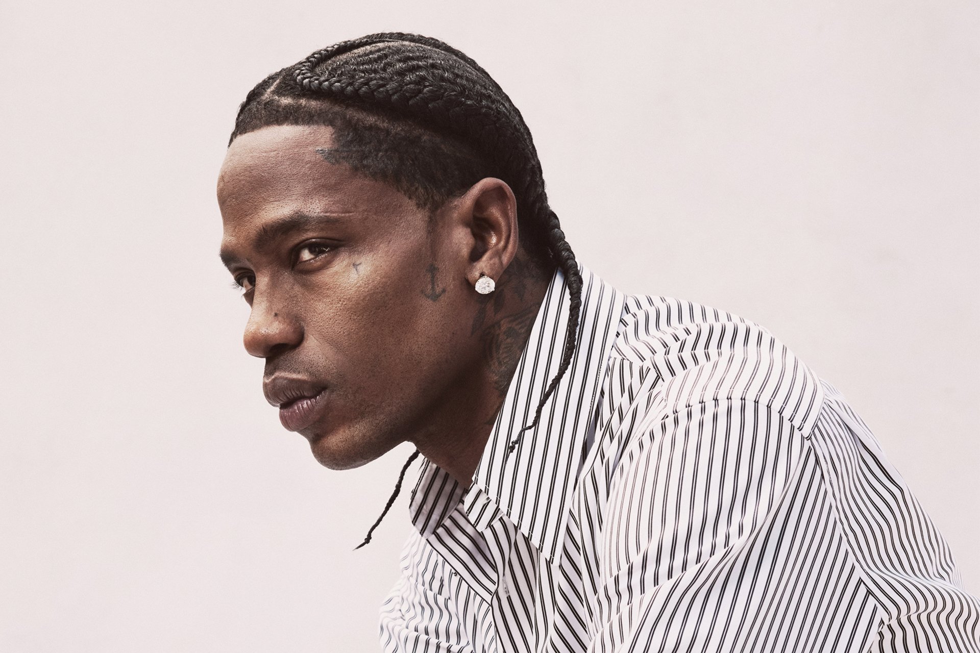 A man with cornrow braids and a diamond earring looks off to the side in a portrait against a plain white background, wearing a black-and-white striped shirt.