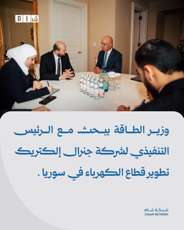 Four people in business attire sit around a white-clothed table with water bottles during a meeting, with Arabic text below and a "Shaam Network" logo.