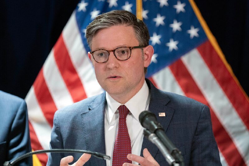A man in a blue suit and red tie stands at a microphone with an American flag behind him.