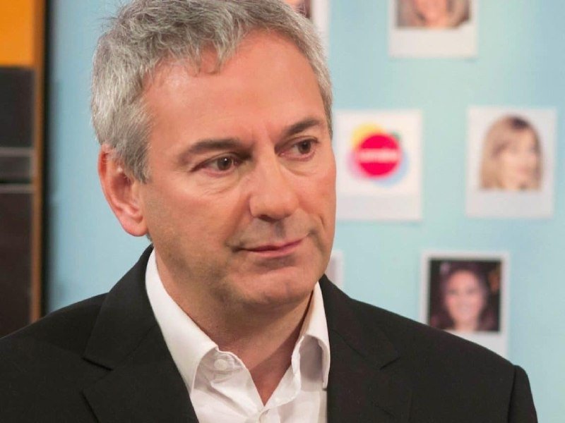 A gray-haired man in a dark suit and white shirt stands before a light blue wall displaying several portrait photographs.