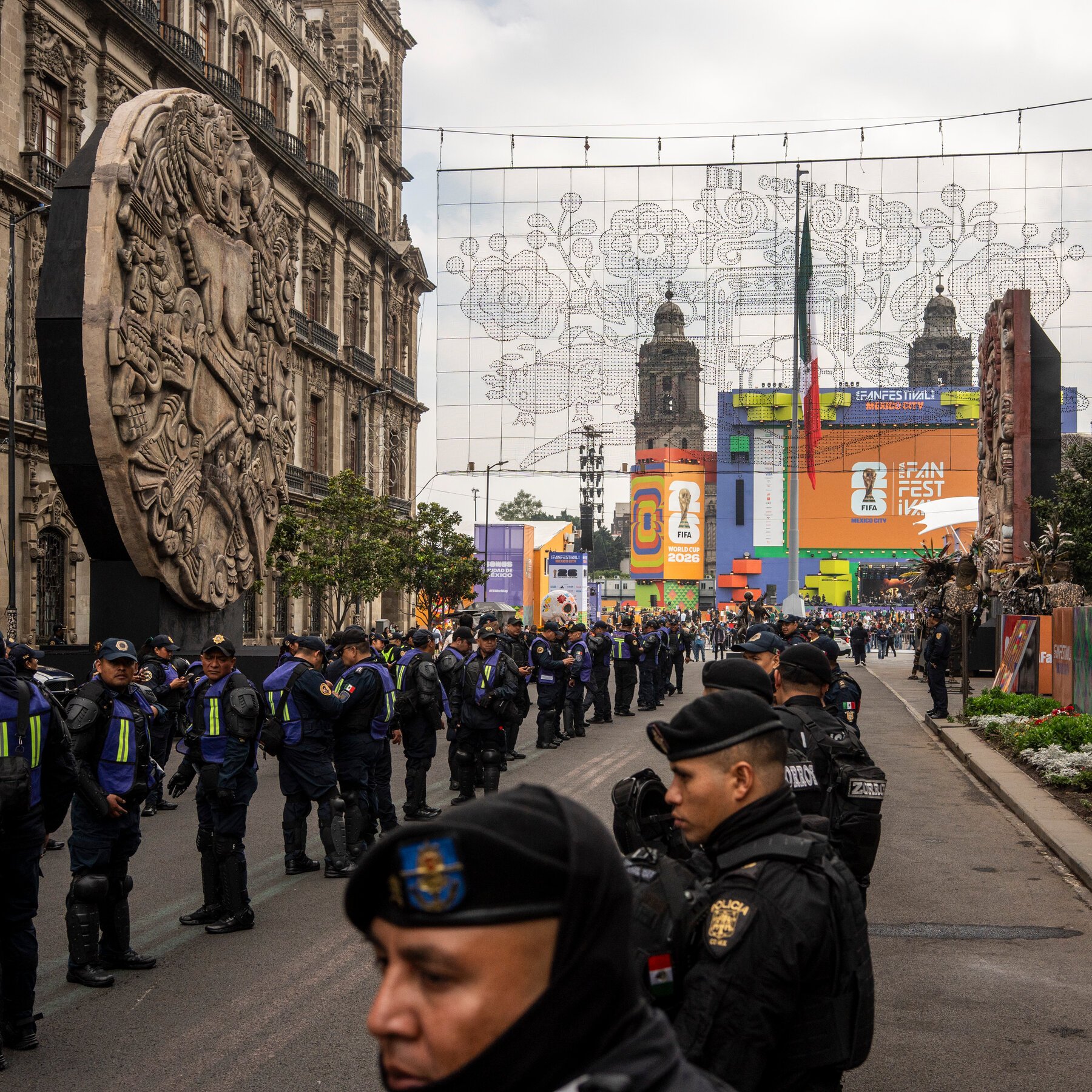 Police officers in dark uniforms stand along a city street near a large stone monument, with colorful "FIFA Fan Festival Mexico City" signage and scaffolding visible in the background.