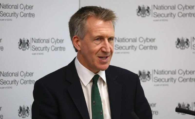 A grey-haired man in a dark suit and green tie speaks while standing in front of a backdrop displaying the National Cyber Security Centre logo.
