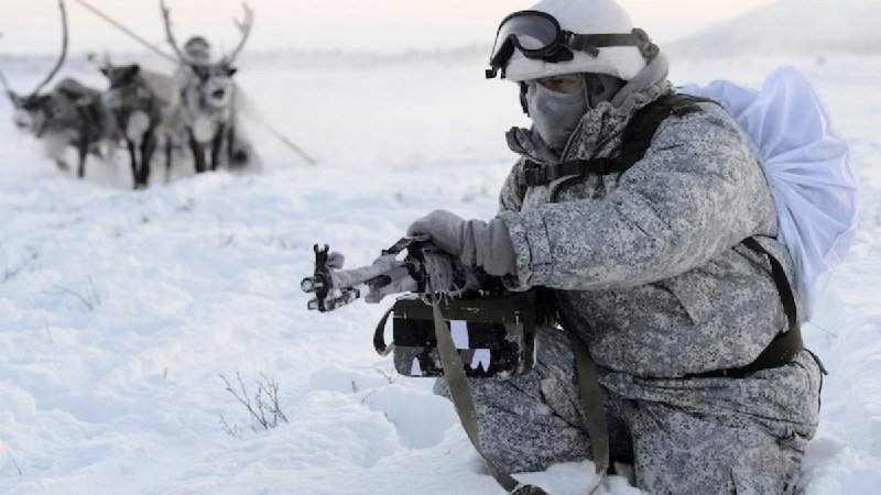 A soldier in white winter camouflage and a face mask crouches in the snow, aiming a rifle, with another person and reindeer visible in the background.