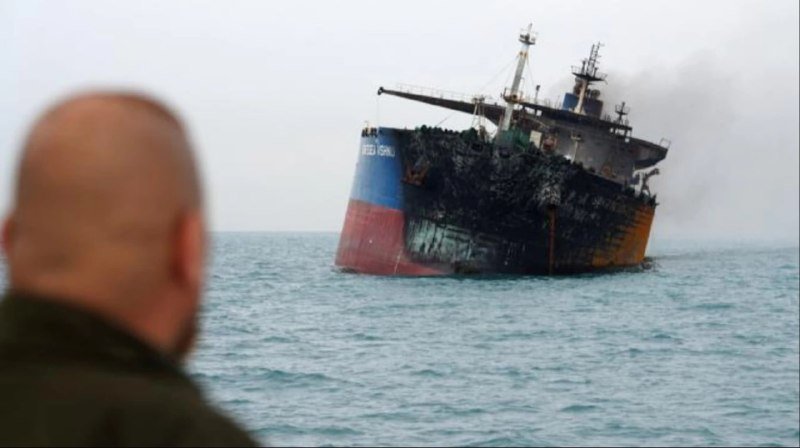 A man with a shaved head watches a blackened, smoke-damaged cargo ship sinking in the ocean.
