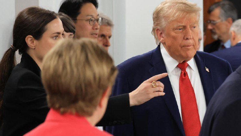 A man in a navy suit and red tie stands among a group of formally dressed people indoors, as a woman gestures toward him with an outstretched hand.