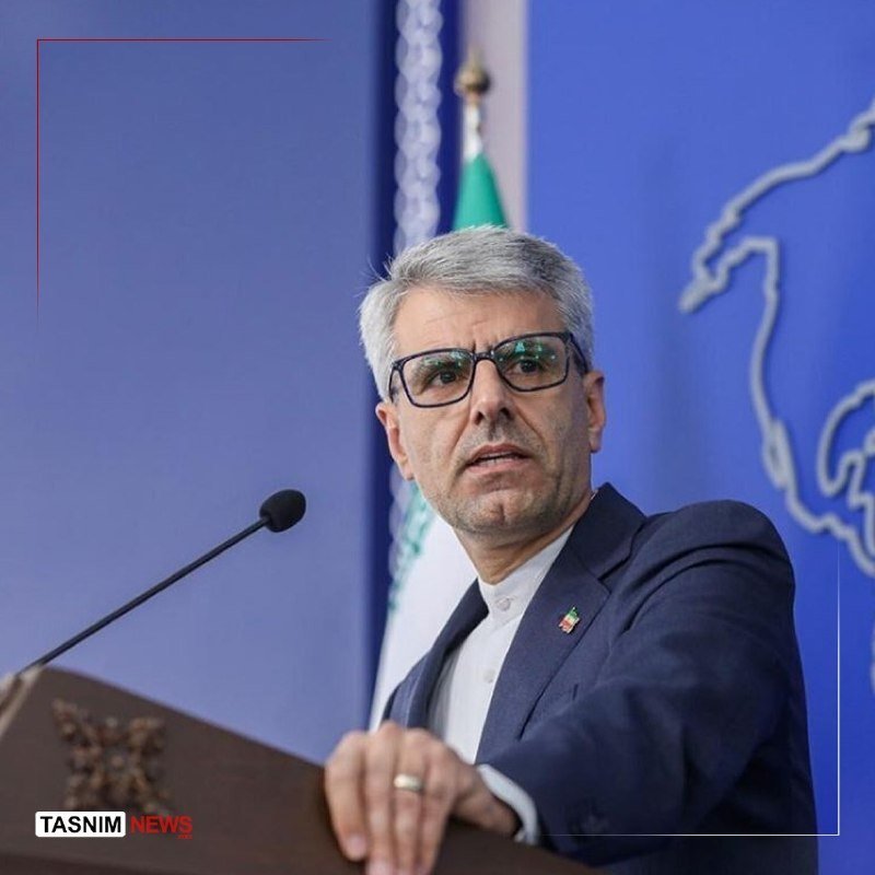 A gray-haired man in glasses and a dark suit speaks at a podium, with a flag and blue backdrop behind him; a "Tasnim News" logo appears in the corner.