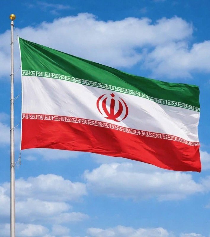 The flag of Iran—featuring green, white, and red horizontal stripes with a red central emblem—waves from a metal flagpole against a partly cloudy blue sky.