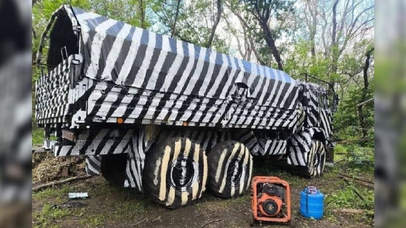 A large camouflage-patterned truck with black and white stripes sits in a wooded area, accompanied by an orange generator and blue fuel container on the ground nearby.