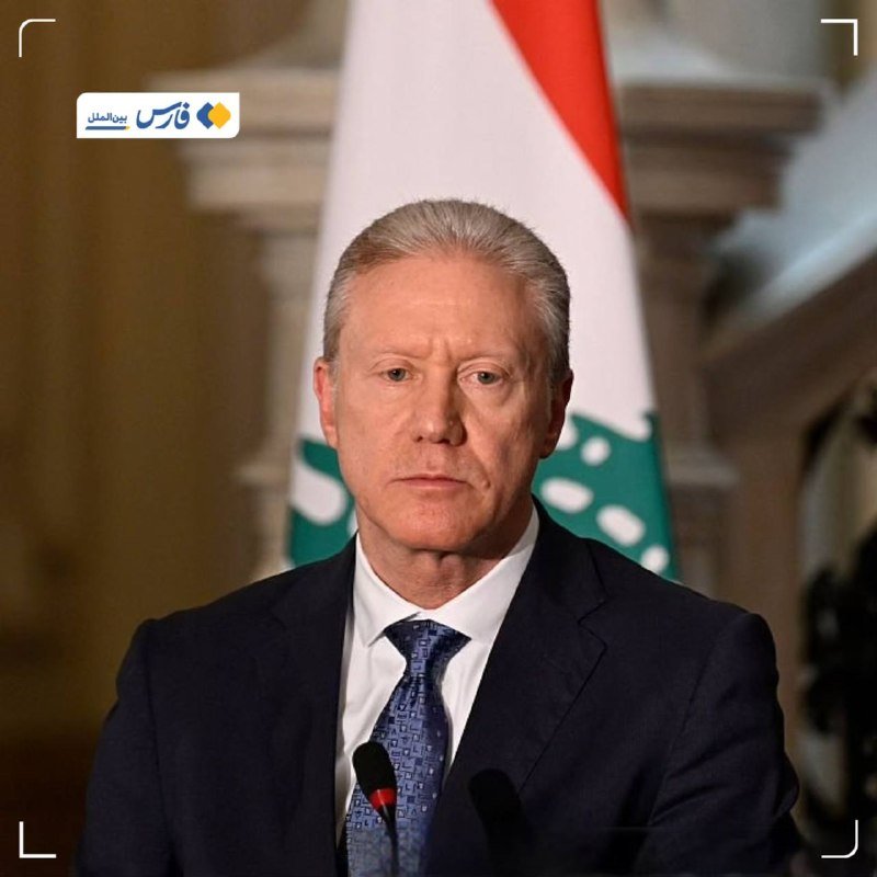 An older man in a dark suit and blue tie stands at microphones in front of a Lebanese flag.