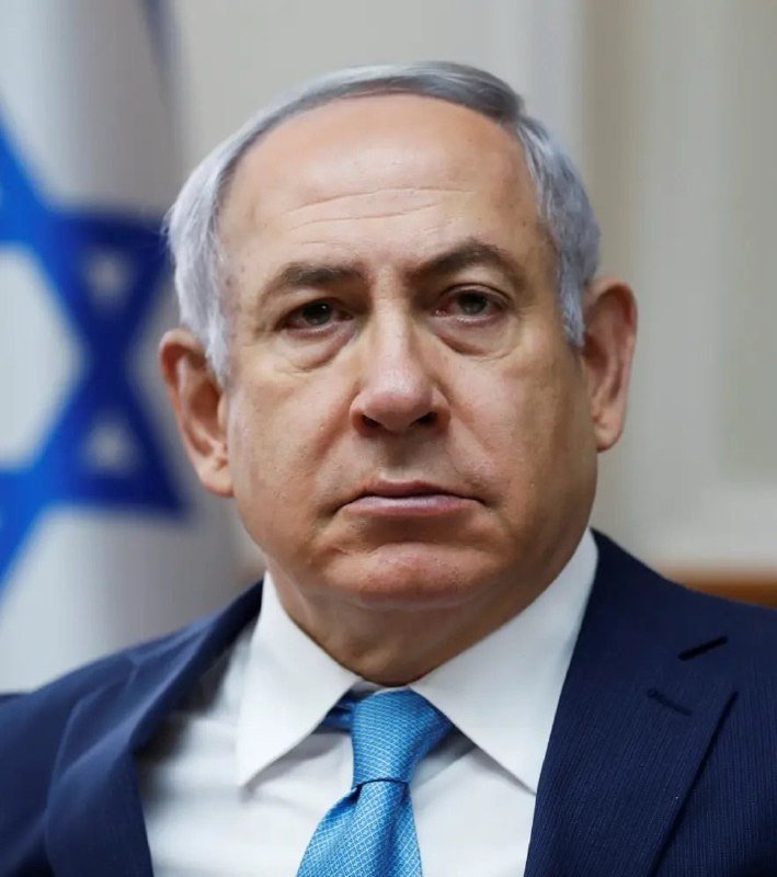 A man in a navy suit and blue tie is pictured in a close-up portrait with an Israeli flag partially visible behind him.