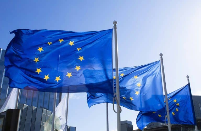 Three European Union flags fly on flagpoles against a clear blue sky in front of a modern glass building.