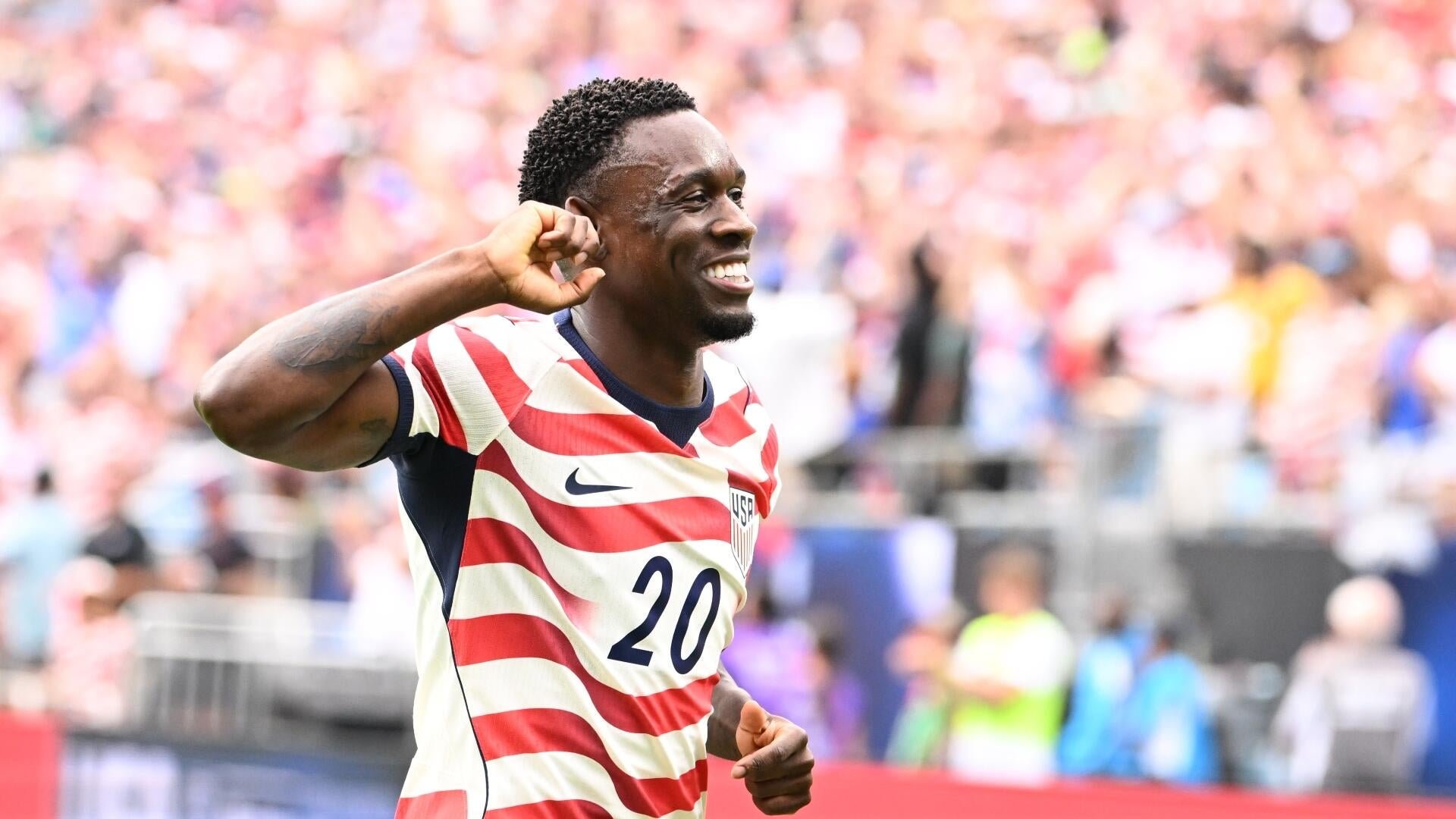 A soccer player wearing a red, white, and blue striped jersey with the number 20 smiles and raises a clenched fist in celebration before a stadium crowd.