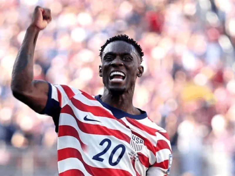 A soccer player wearing a red and white striped USA jersey with the number 20 raises his fist in celebration before a stadium crowd.